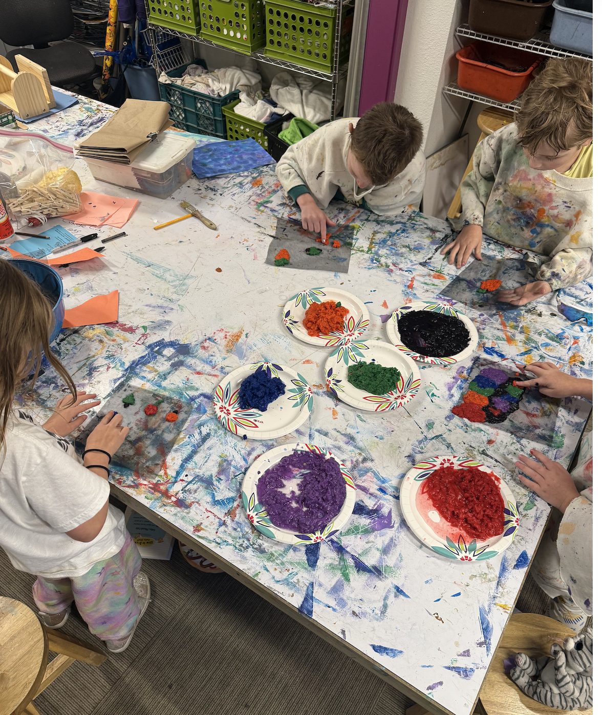 Children making colorful textured art at a paint-splattered communal table